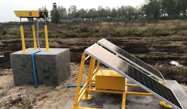 Solar powered field monitoring station with angled solar panels on a yellow frame beside a concrete pedestal with a mounted sensor array in a dirt field