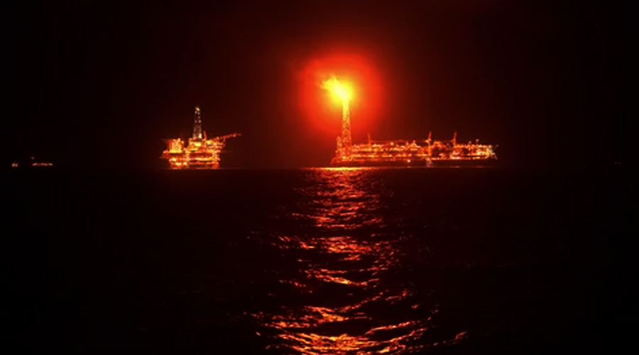 Offshore oil rig at night with a bright flare stack and reflections on the dark water surface
