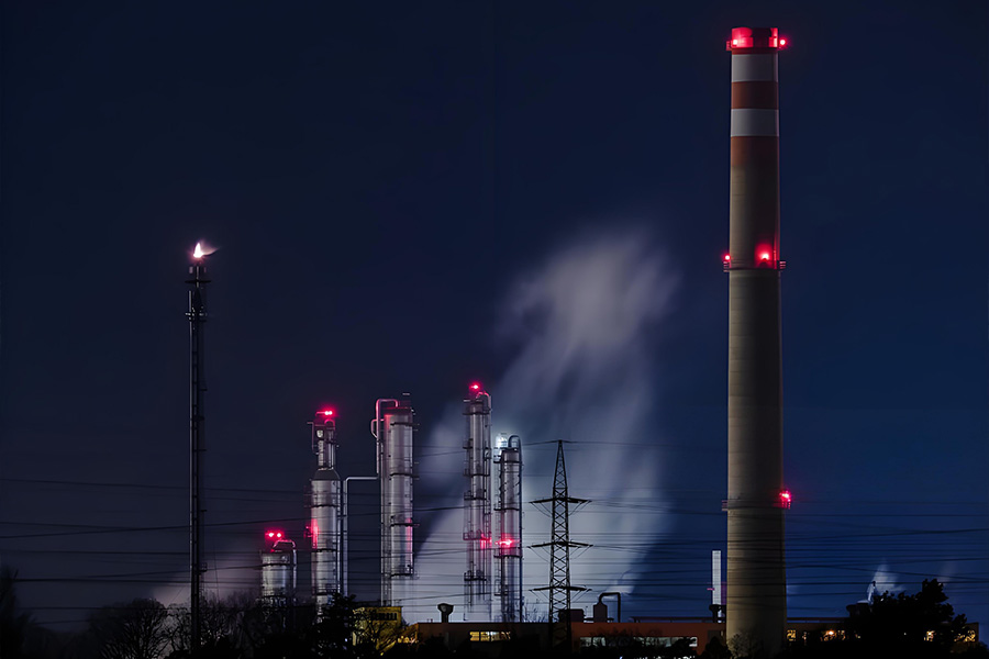 Night view of an industrial plant with a tall smokestack emitting steam and red warning lights on the towers.