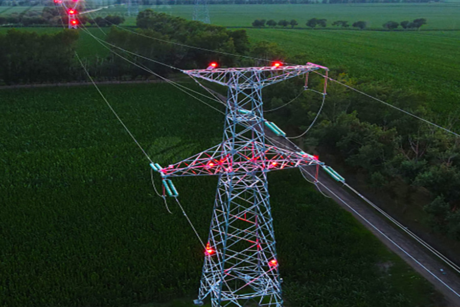 High-voltage transmission tower with multiple wires and red warning lights over a green rural field and a road nearby.