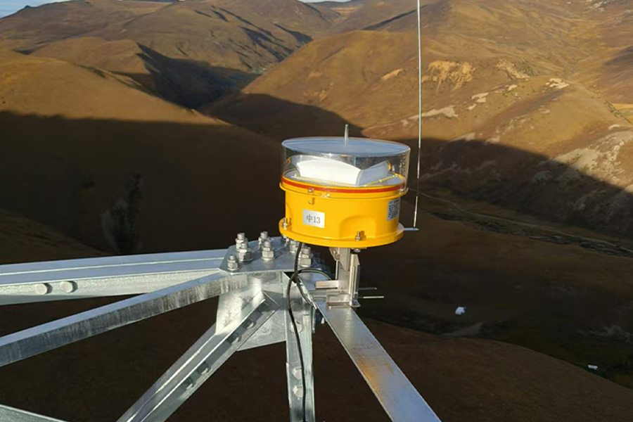 Yellow round weathermonitoring sensor with a clear dome mounted on a metal lattice tower against brown hilly landscape in the background