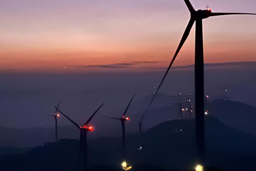 Wind turbines on a hill at sunset with silhouettes against a purple-orange sky and red aircraft lights on the towers/rotors.