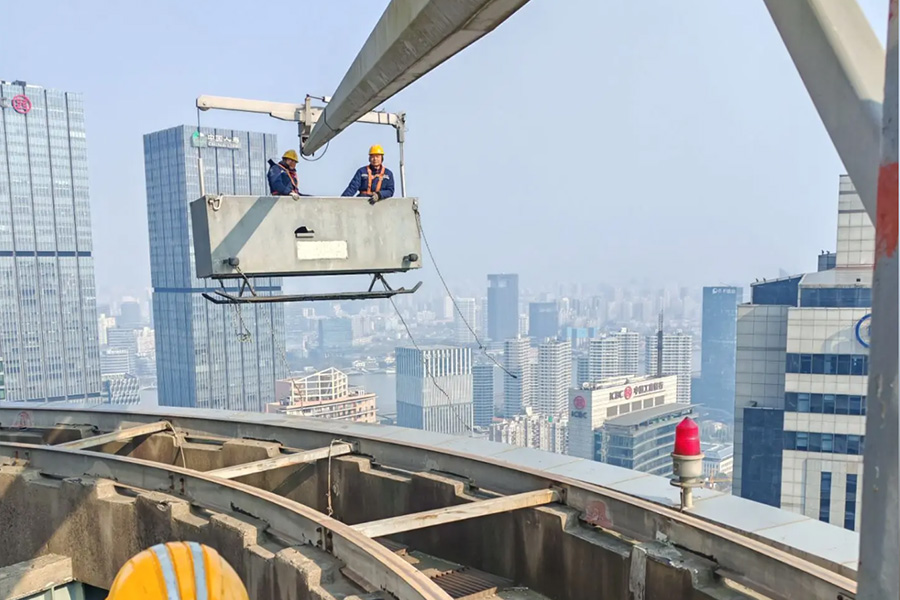 Two construction workers wearing hard hats stand on a suspended work platform above a city skyline, being lifted by a crane over a curved rooftop track.