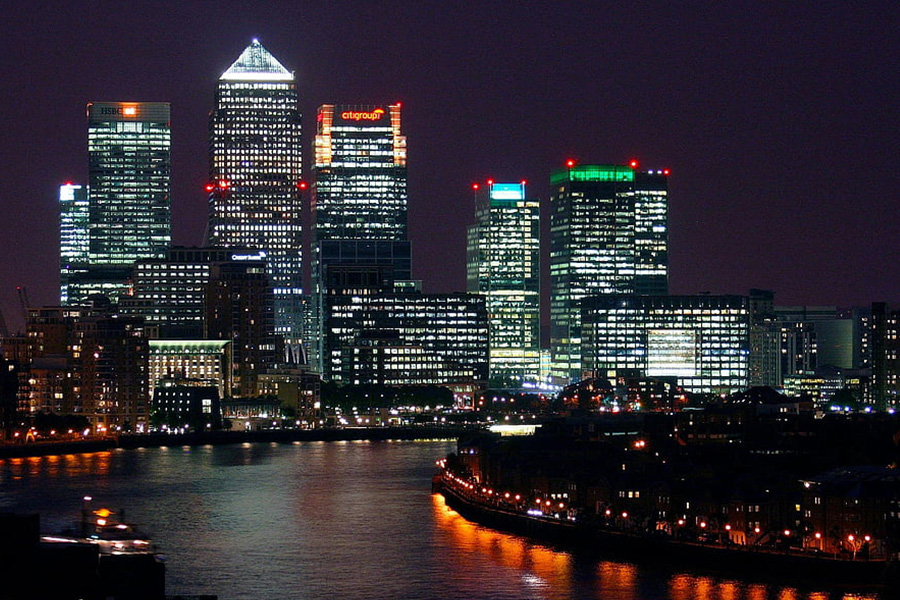 Nighttime city skyline along a calm river with illuminated skyscrapers reflecting in the water