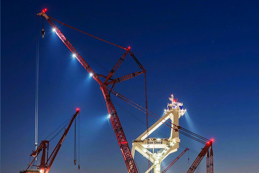 Nighttime harbor construction site with tall red lattice cranes and bright work lights against a dark blue sky