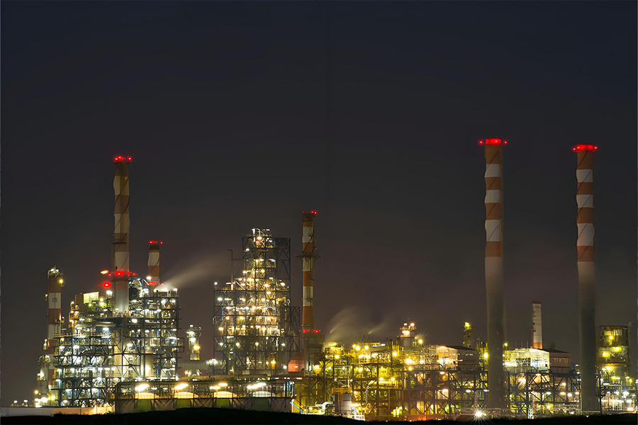 Night view of a lit industrial refinery with tall smokestacks and complex piping at night