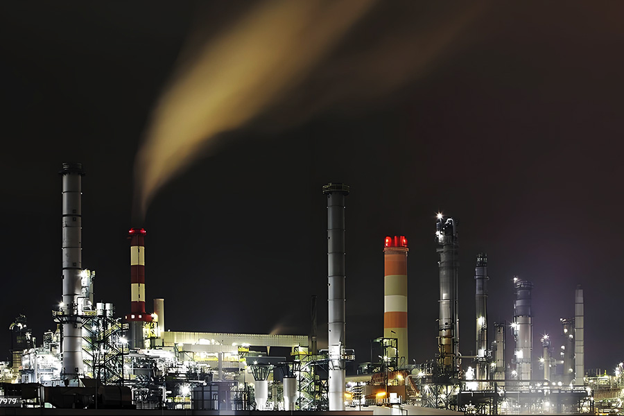 Nighttime view of a large oil refinery with multiple lit smokestacks and visible vapor plumes on a dark sky