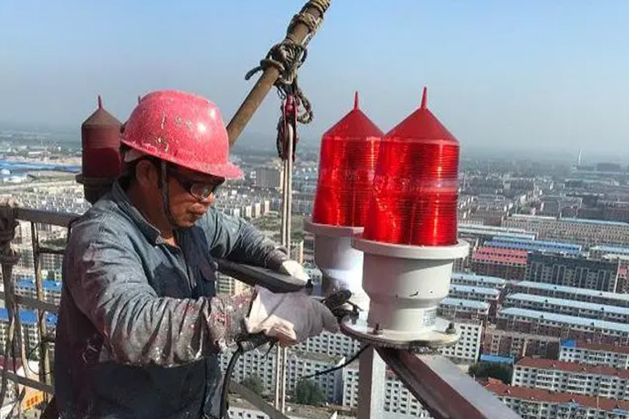 Worker in a red hard hat and gloves installing a red warning beacon atop a rooftop tower, city skyline in background