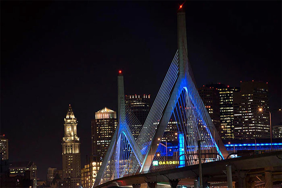 Night view of a cable-stayed bridge illuminated in blue at night with a lit city skyline in the background, including tall office towers and the TD Garden area nearby.