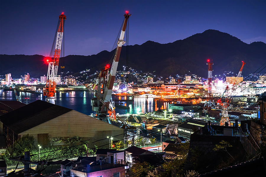 Nighttime port scene with large cranes and an illuminated harbor, mountains in the background