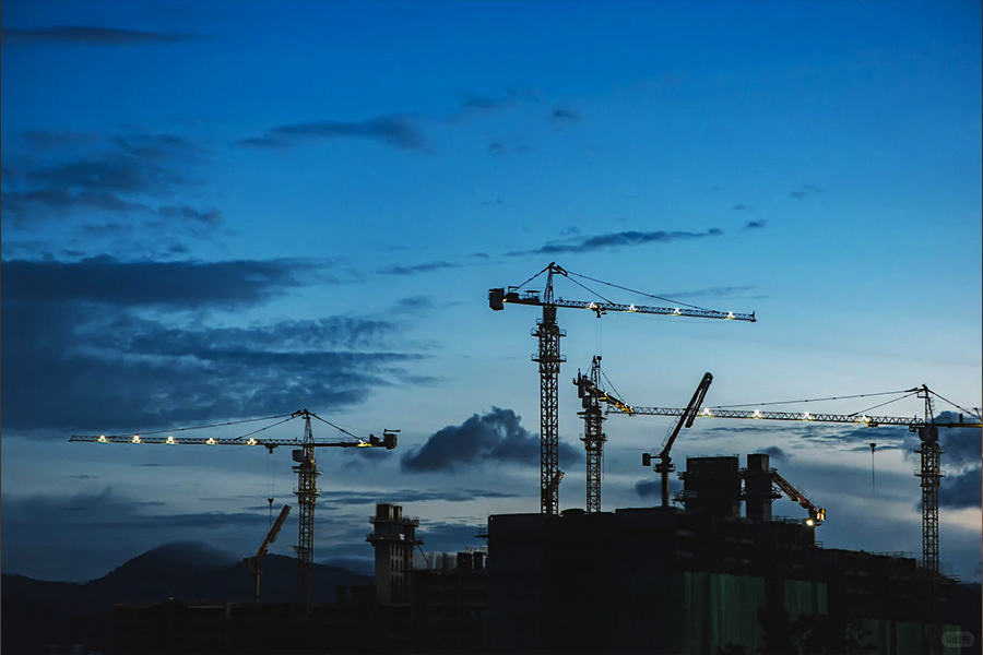 Construction site at dusk with several cranes against a blue sky, building framework in silhouette.