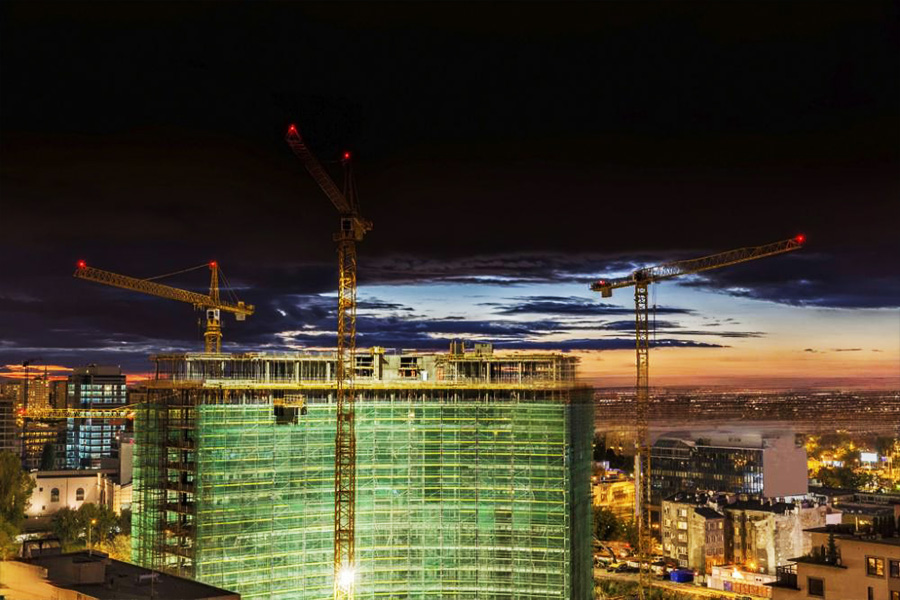 Night view of a construction site with an illuminated building under scaffolding and two cranes towering above.