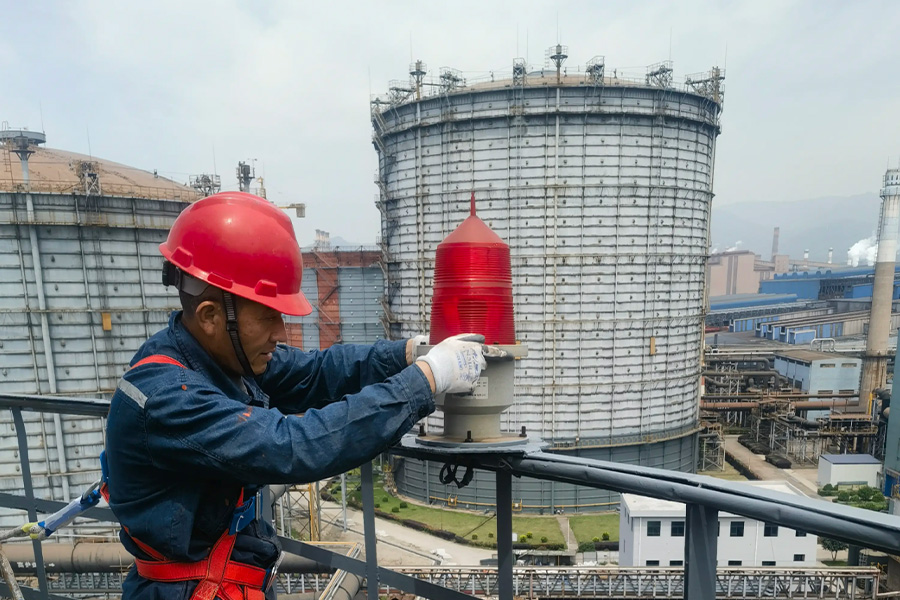 Industrial worker in a red hard hat and safety harness adjusts a red beacon on a railing with large storage tanks in the background