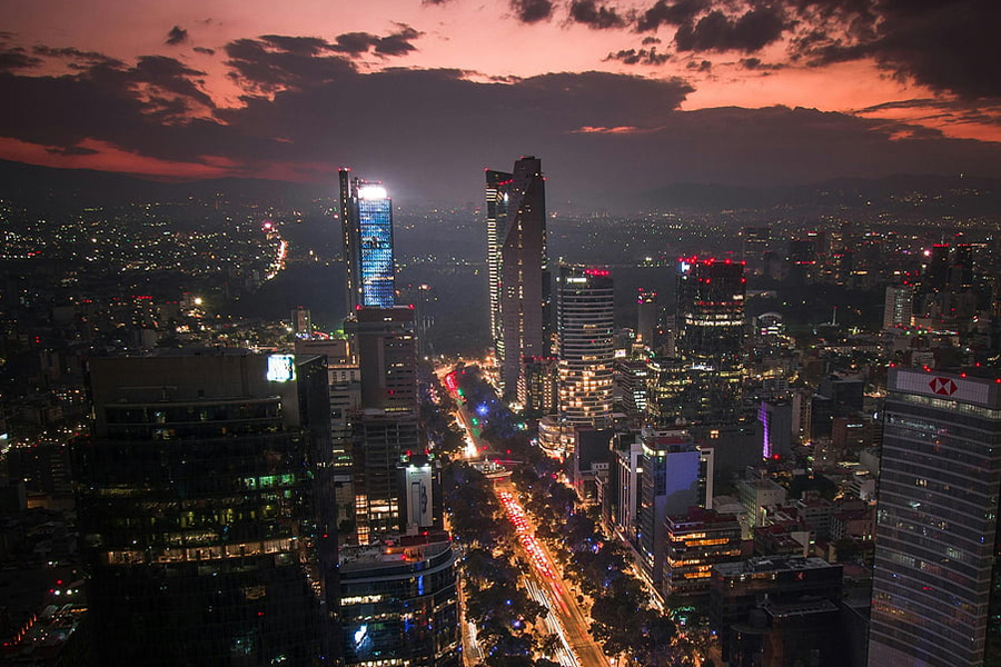 Aerial night cityscape with tall skyscrapers, bright streetlights along a central avenue, and a red-orange sunset sky above the horizon.