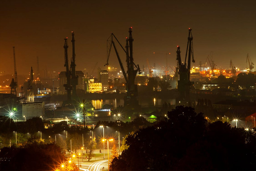 Nighttime port scene with tall cranes silhouetted against a hazy orange sky and bright harbor lights below in the foreground